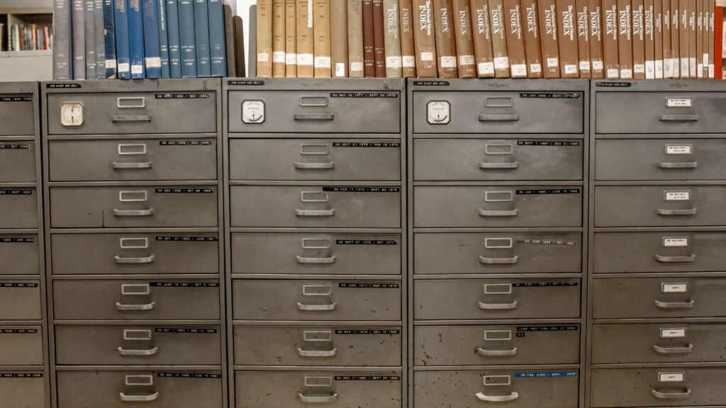 Three rows of gray metal filing cabinets are shown, with books and binders stored on top of them—classic tools before business file sharing made it easier to share files in business. Each row has a clock affixed above the drawers.