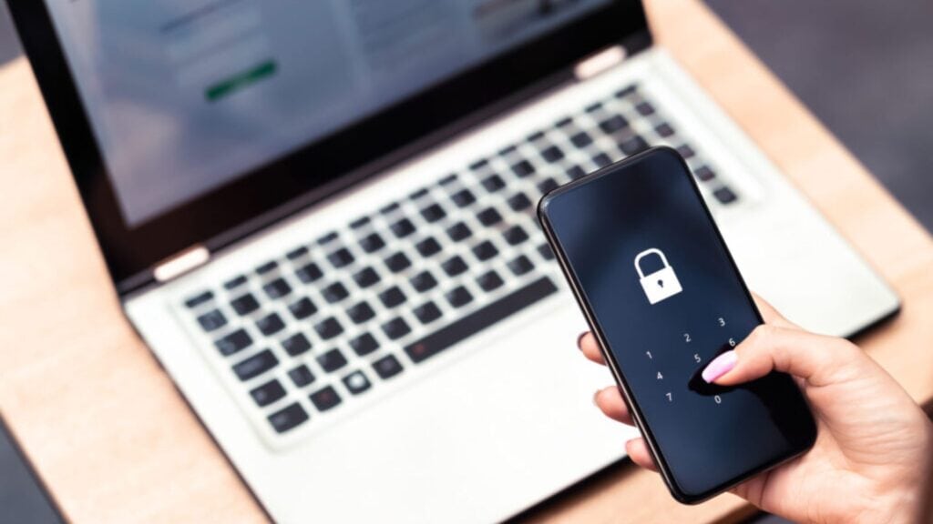 A person holds a smartphone displaying a locked padlock icon and a number pad, in front of an open laptop on a wooden desk, highlighting the importance of secure file sharing in today's digital world.