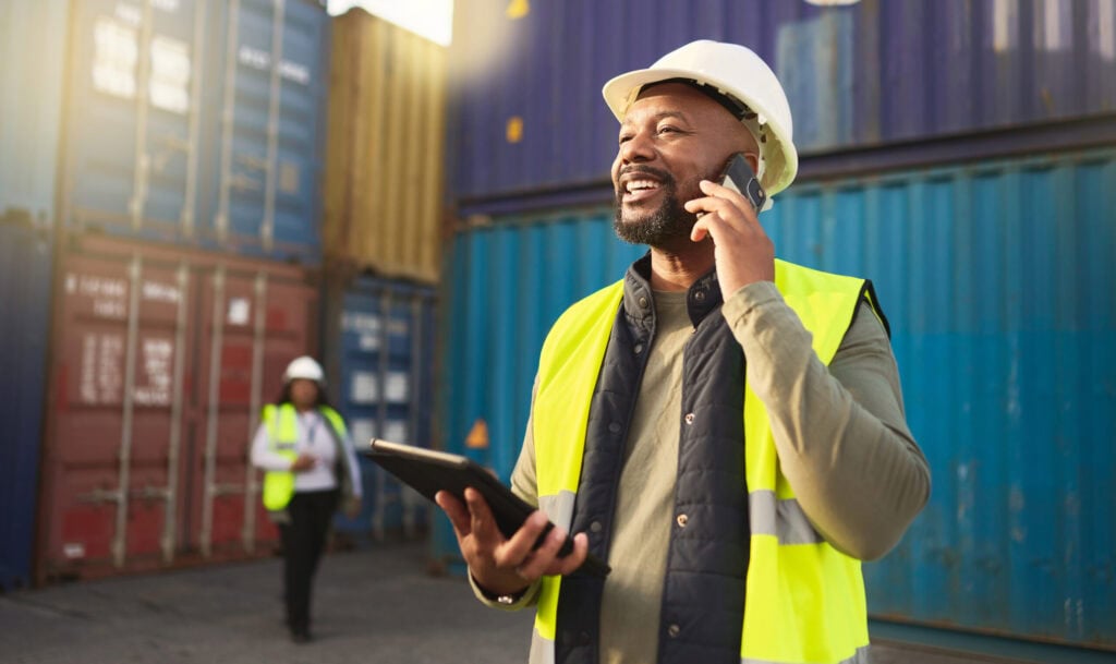Man wearing a hard hat and safety vest talks on the phone and holds a tablet at a shipping container yard, emphasizing the importance of cyber security in construction; another worker is visible in the background.