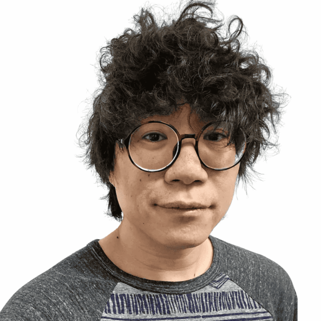 A person with curly dark hair, round glasses, and a grey and blue shirt is looking at the camera against a plain white background.