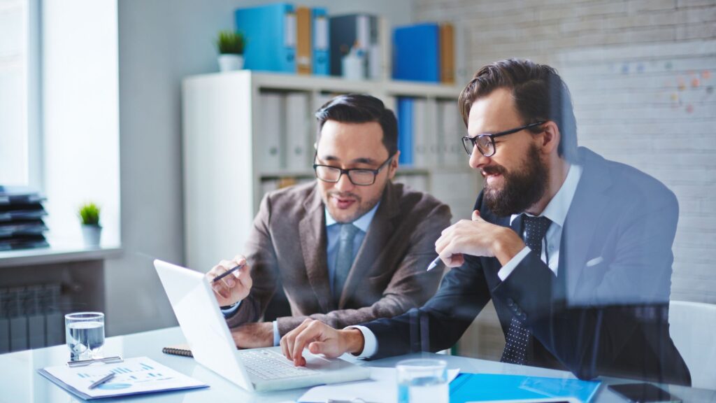 Two men in business attire sit at a desk, looking at a laptop screen and discussing documents—collaborating with a good IT consultant in a modern office setting.