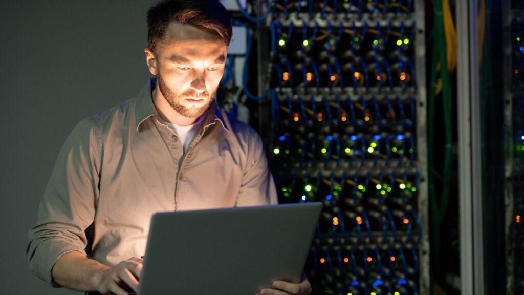 A person working on a laptop in a server room, leveraging IT Service Management principles amidst illuminated servers and network equipment in the background.