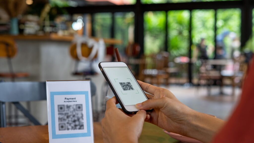 A person scans a QR code for payment using a smartphone at a table in a café, highlighting the importance of cybersecurity and how to protect yourself from threats like Quishing.