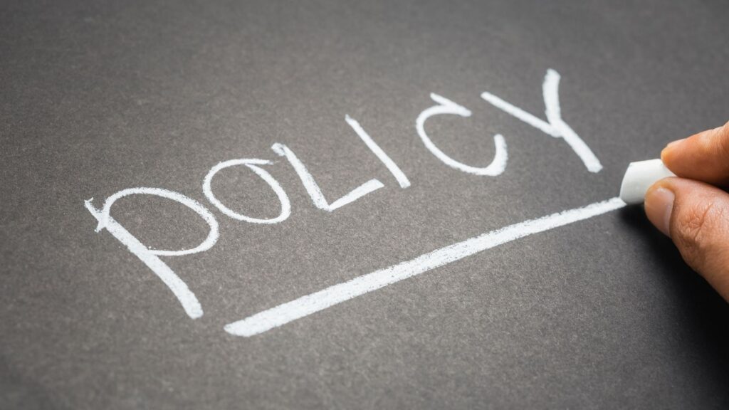 The word "policy" is written in white chalk on a blackboard, with a hand holding a piece of chalk and drawing a line underneath—symbolizing the clarity of an access control or Conditional Access Policy.