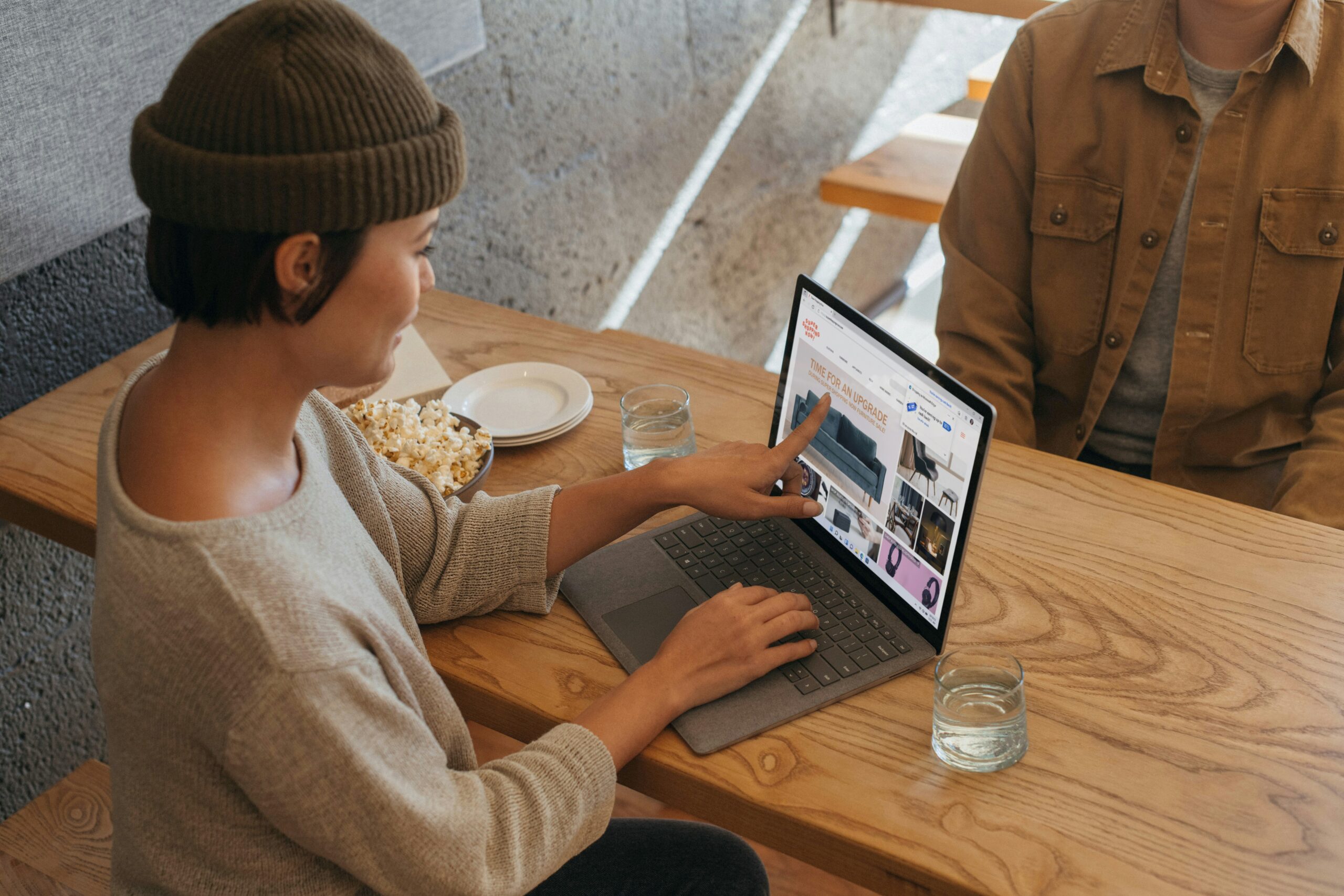A person wearing a beanie and sweater on a laptop in a cafe setting, pointing at the screen, looking up what is Office 365 and what is it used for.