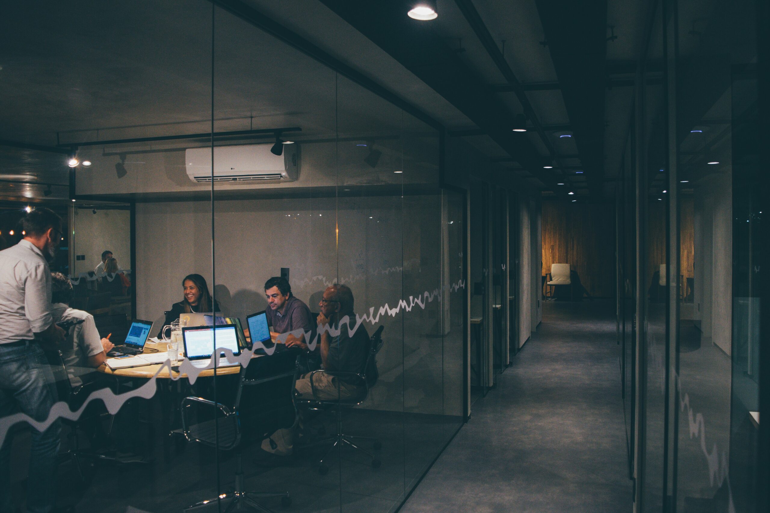 People sitting around a table with laptops and papers in a glass-walled conference room, possibly discussing, what is Office 365 and what is it used for.