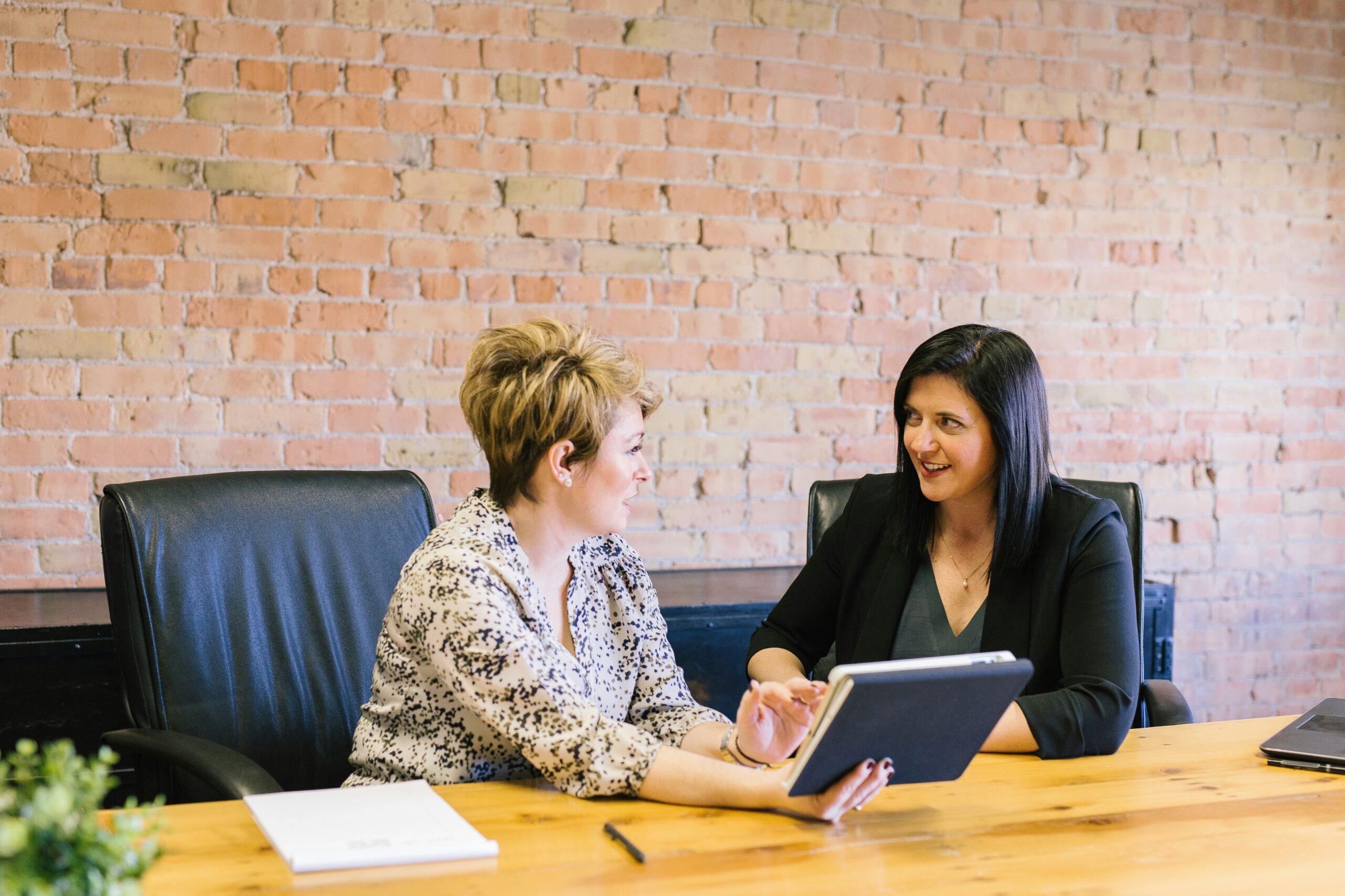 Two women sitting at a wooden table in an office setting with one holding a tablet, engaged in discussion about Office 365 use.